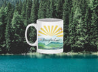 White coffee mug with sunrise rays and mountain landscape graphic, side view