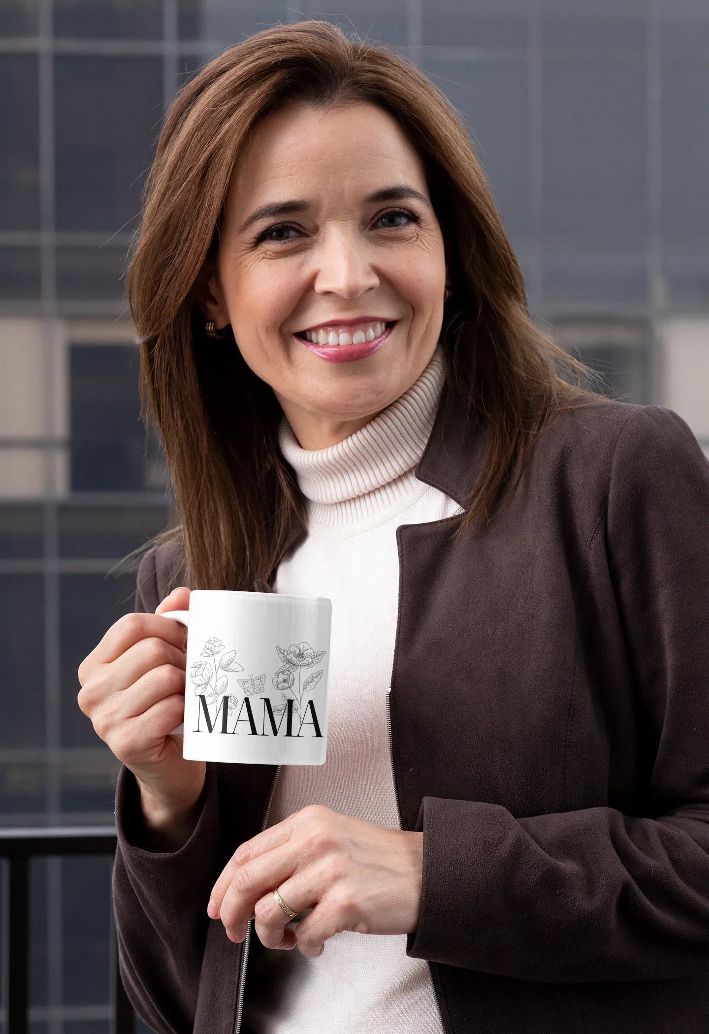 White ceramic coffee mug with 'MAMA' text held by woman in dark jacket