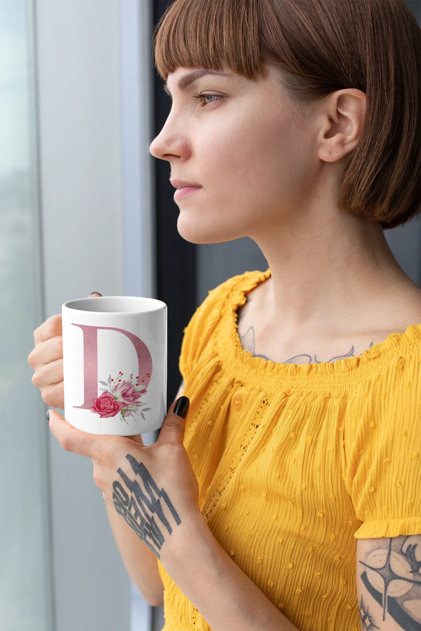White mug with floral letter D design held by a woman in a yellow top.