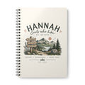 A spiral-bound notebook with a cream-colored cover featuring a mountain landscape illustration and the text 'HANNAH Girls who hike GO FURTHER'.