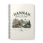 A spiral-bound notebook with a cream-colored cover featuring a mountain landscape illustration and the text 'HANNAH Girls who hike GO FURTHER'.