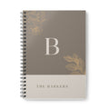 A5 notebook with taupe cover, gold monogram B, and floral corner accents; spiral binding.