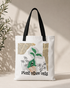 A white canvas tote bag with black shoulder straps, featuring a plant-themed graphic print with the text 'Plant vibes only' and a green sprout illustration, set against a warm beige background with soft leaf shadows.