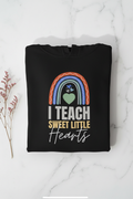 Black unisex hoodie with front graphic: rainbow heart and text “I TEACH SWEET LITTLE Hearts”