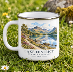 A white enamel-style mug with a silver rim sits on grass outdoors. The mug features a scenic printed landscape of a lake and mountains with the text 'LAKE DISTRICT CUMBRIA • ENGLAND 2026'.