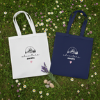 White and navy cotton tote bags on grass, each with mountain sketch and pink heart