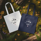 White and navy cotton tote bags on forest floor, each with mountain sketch and pink heart