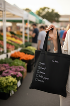Black tote bag held up with 'to buy' checklist: milk, bread, cheese, toilet paper, snacks.