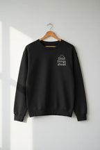 Black crewneck sweatshirt with small chest logo, hanging on a wooden hanger