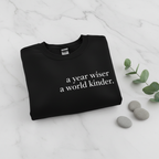 Black sweatshirt with “a year wiser, a world kinder.” text printed on the front.