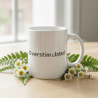 White ceramic mug with handle on table, “Overstimulated.” printed on front