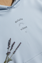 Light blue hoodie with 'delulu vibes' print, small heart, and lavender-like floral stems.