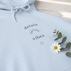 Light blue hoodie with 'delulu vibes' print, heart icon, leafy flower graphic, chest close-up.