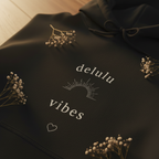 Black hoodie with 'delulu vibes' text graphic, heart and floral motif, front view.