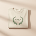Cream personalized name T-shirt with green floral wreath monogram reading “EMMA est. 2019”