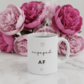 White ceramic engagement mug on marble with pink floral backdrop; "engaged AF" and hearts