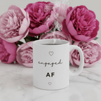 White ceramic engagement mug on marble with pink floral backdrop; "engaged AF" and hearts