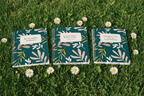 Three teal A5 spiral notebooks with botanical covers on green grass, label visible
