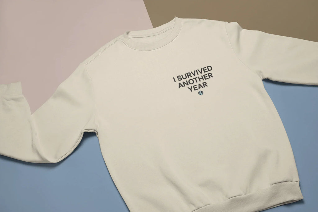 Light cream crewneck sweatshirt with small chest text graphic “I SURVIVED ANOTHER YEAR”