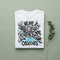 White unisex T-shirt with “We Are God’s Creatures” animal graphic, flat lay.