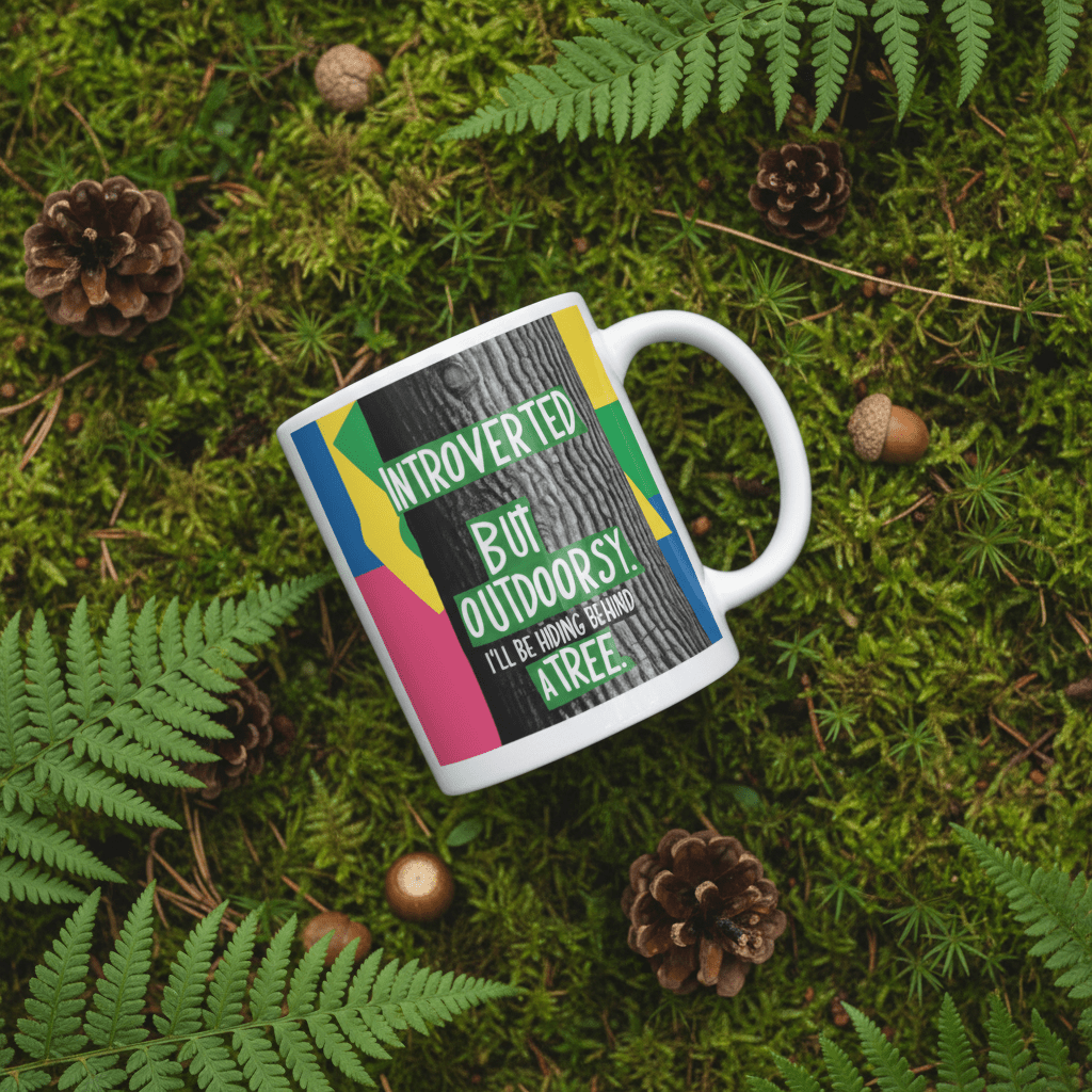 White ceramic mug with green “Introverted” and “Outdoorsy” text graphic on outdoor scene.