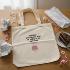 Cream canvas tote bag with birthday quote and pink cake graphic, laid on a table