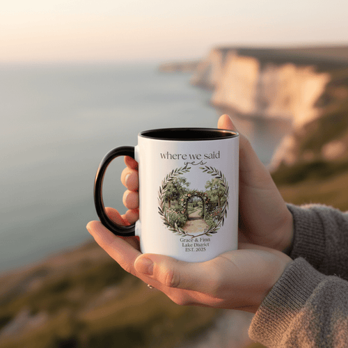 White accent mug with black handle held near ocean view, "where we said yes" floral arch