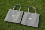 Pair of light grey cotton tote bags with text “BFF.” and “AVA”.