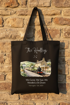 Black canvas tote bag displayed on a brick wall, featuring a printed train and cottage scene.
