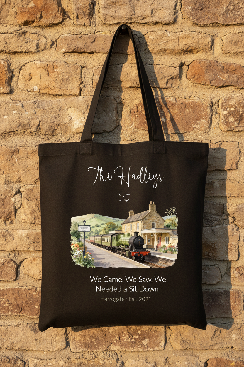Black canvas tote bag displayed on a brick wall, featuring a printed train and cottage scene.