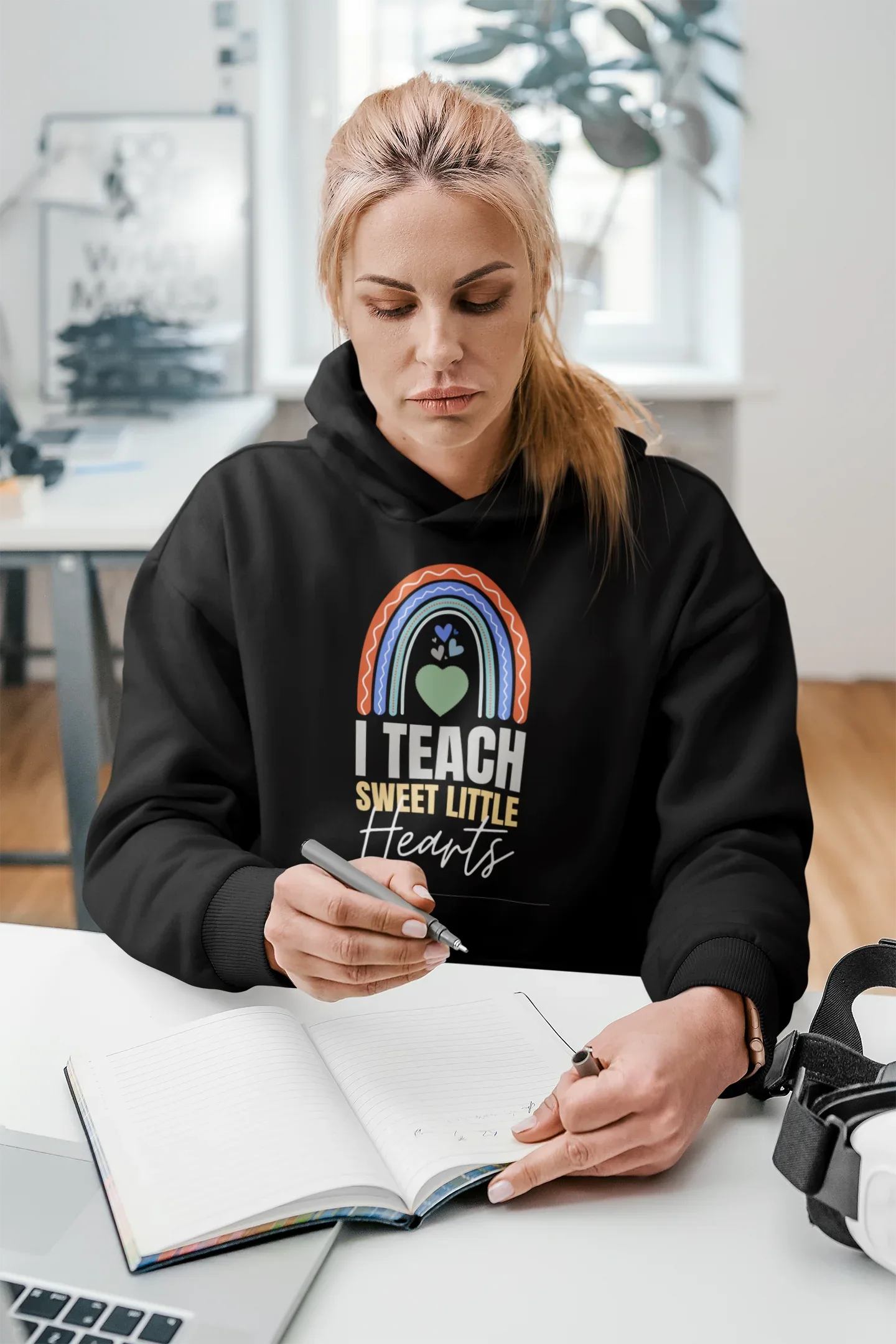 Black unisex hoodie worn by person, featuring front rainbow heart graphic and “I TEACH SWEET LITTLE Hearts”