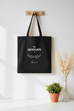 Black graduation tote bag hanging on wall with “THE GRADUATE” and name Grace