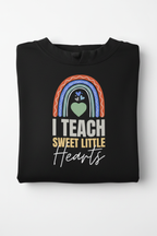 Black unisex pullover hoodie with large front print: rainbow arch, heart icon, “I TEACH”