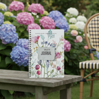 Isla's personalised Bloom Where You Are Planted A5 floral journal standing upright on a wooden garden bench in front of hydrangeas in a British country garden