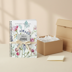 Nell's personalised Bloom Where You Are Planted A5 floral journal standing upright with a cream ribbon tied around it beside an open gift box with tissue paper, a kraft gift bag and a folded card on a cream linen surface