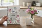 Esme's personalised Bloom Where You Are Planted A5 floral journal standing upright on a wooden kitchen table held by a hand, with a mug of tea and a vase of garden roses in a bright British kitchen