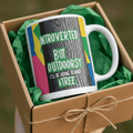 White mug in a gift box, showing “Introverted but Outdoorsy” colorful print.