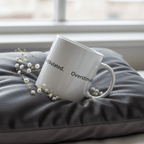 White ceramic mug tilted on dark fabric, printed text “overstimulated.”