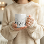 White ceramic coffee mug with 'MAMA' text held by hands in a knit sweater