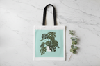 White tote bag with black handles and green monstera leaf graphic on front