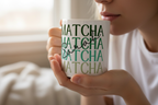 White ceramic mug with repeating green MATCHA text, held near a person’s face.