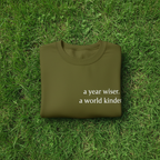 Olive sweatshirt with “a year wiser, a world kinder.” text on the front.