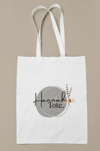 White cotton tote bag with circular “Hannah’s Tote” print and small botanical sprig