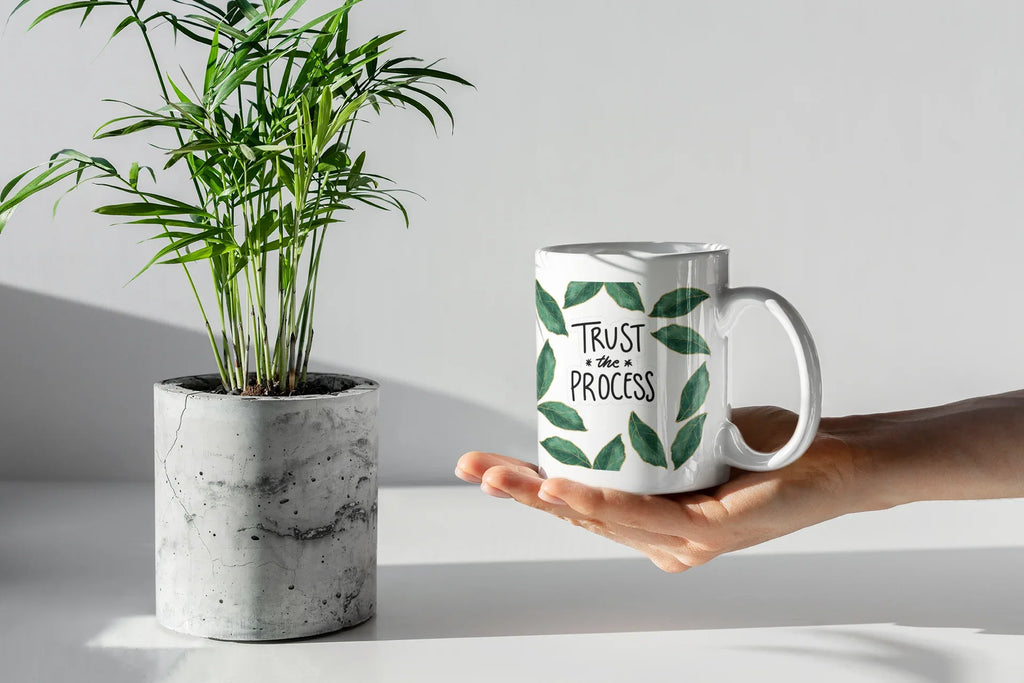 White coffee mug with green leaf wreath design and “Trust the Process” in an open hand