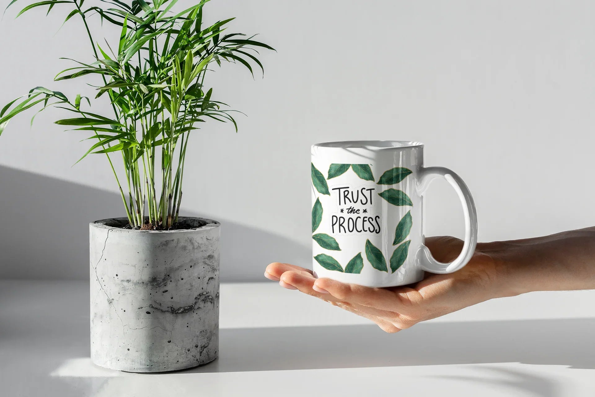 White coffee mug with green leaf wreath design and “Trust the Process” in an open hand