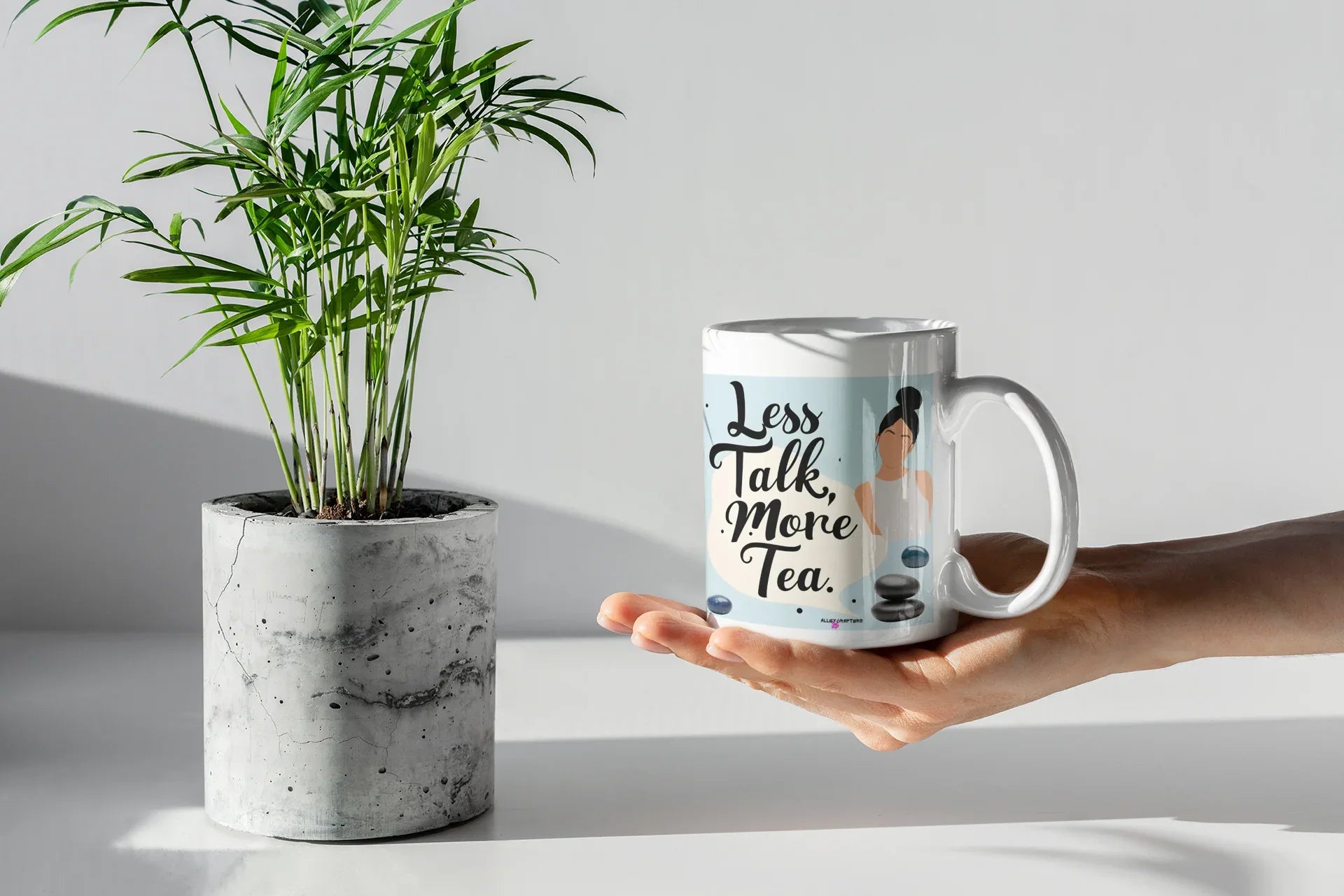 Hand holding a white tea mug with 'Less Talk, More Tea' text and stone graphic