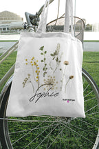 White canvas tote bag with botanical plants print and cursive logo, short handles
