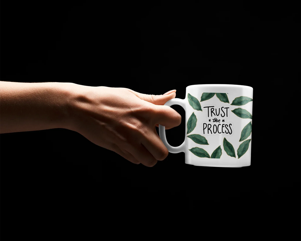 White mug with green leaves and “Trust the Process” text as a hand grips the handle