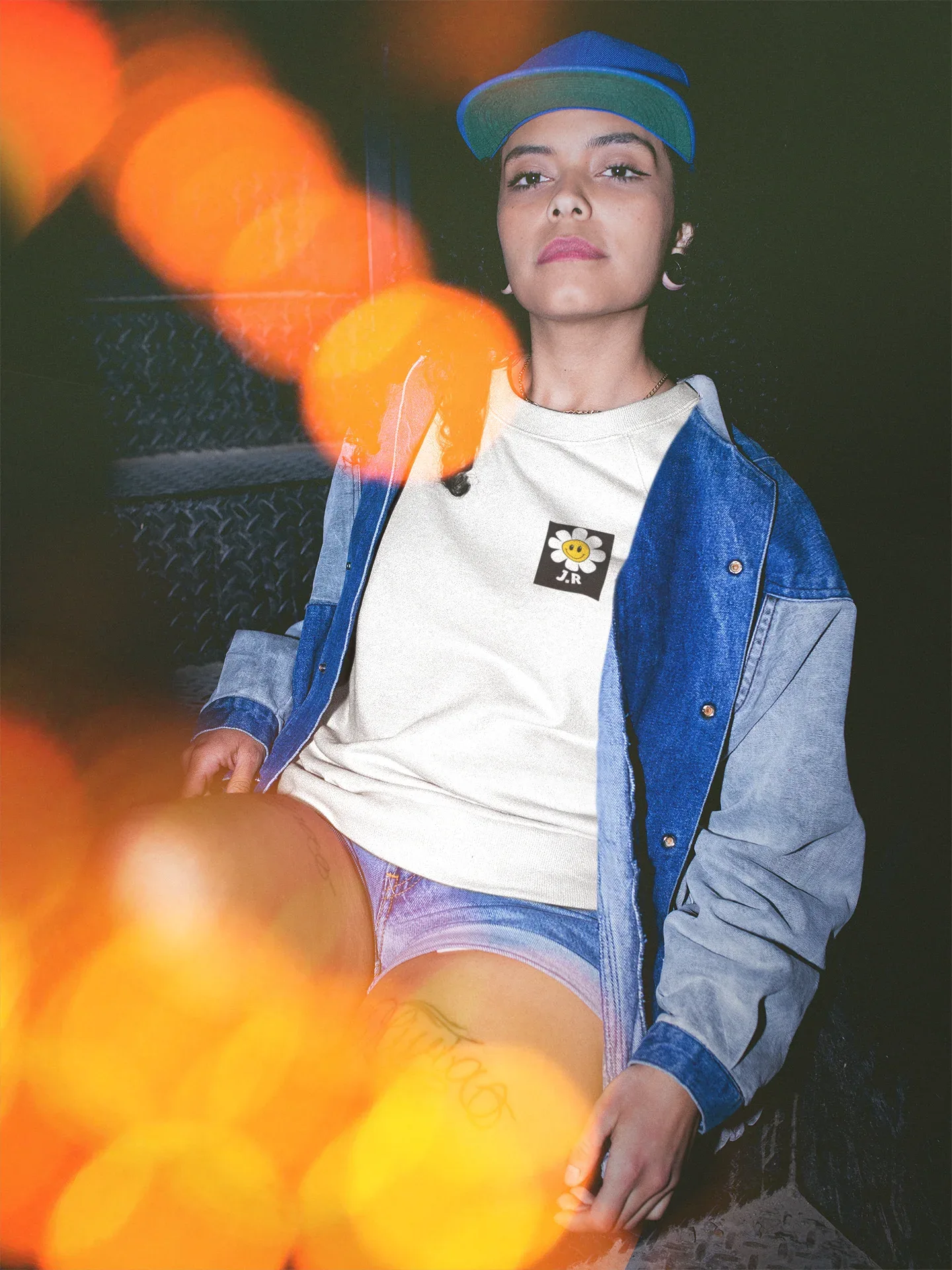 White crewneck sweatshirt with flower smiley patch on chest, worn under denim.