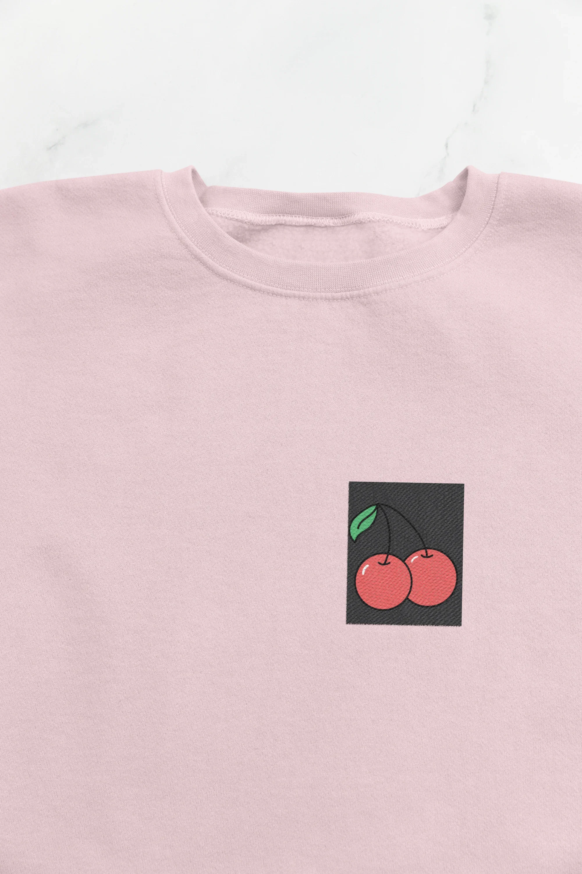 Light pink crewneck sweatshirt close-up with cherry embroidery patch on chest